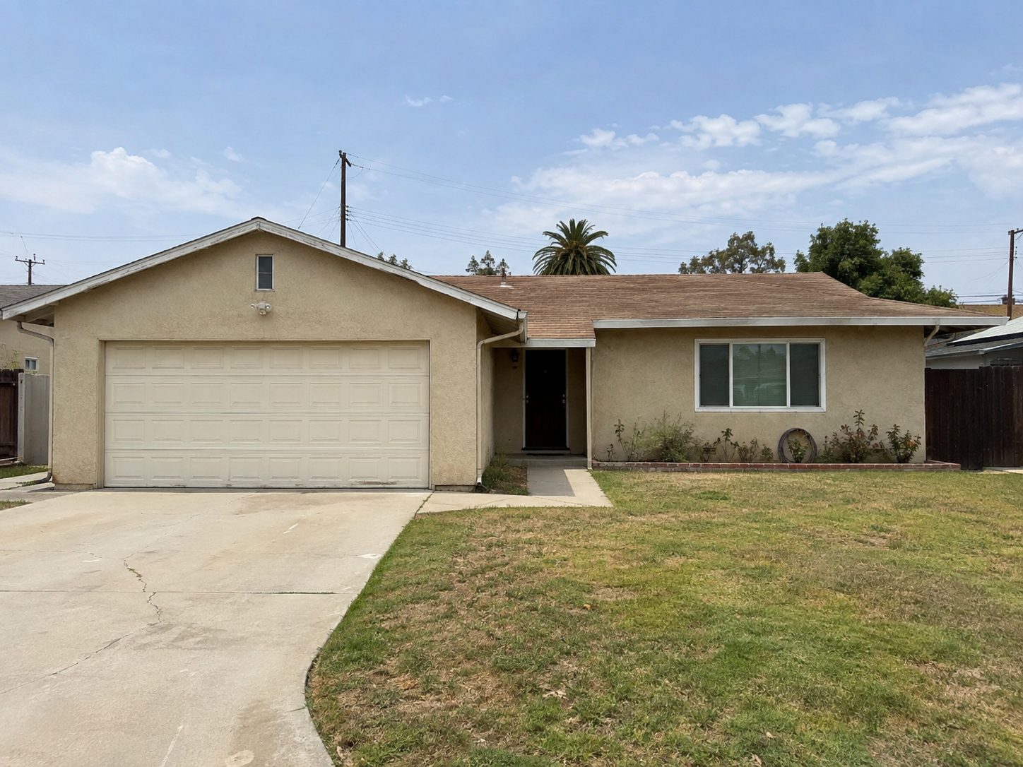 Beige stucco ranch house with a cream garage door, patchy grass, and a cracked concrete driveway