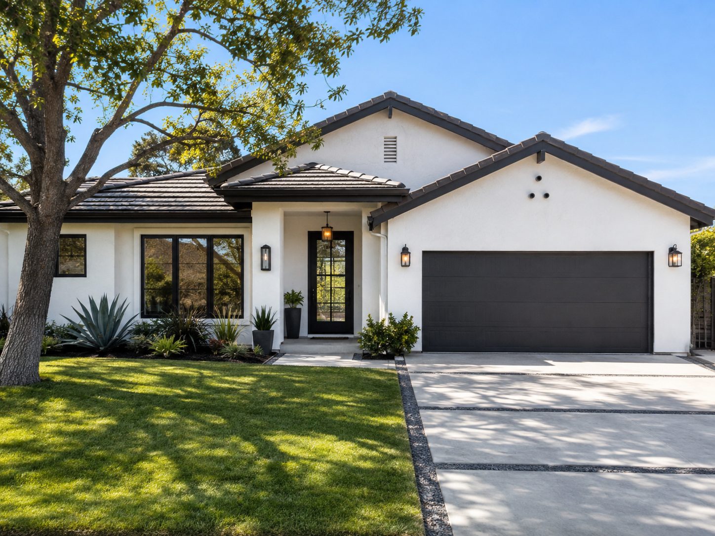 Single story white stucco house with a dark tile roof, black framed windows, a dark garage door, drought tolerant planting, and a wide concrete driveway