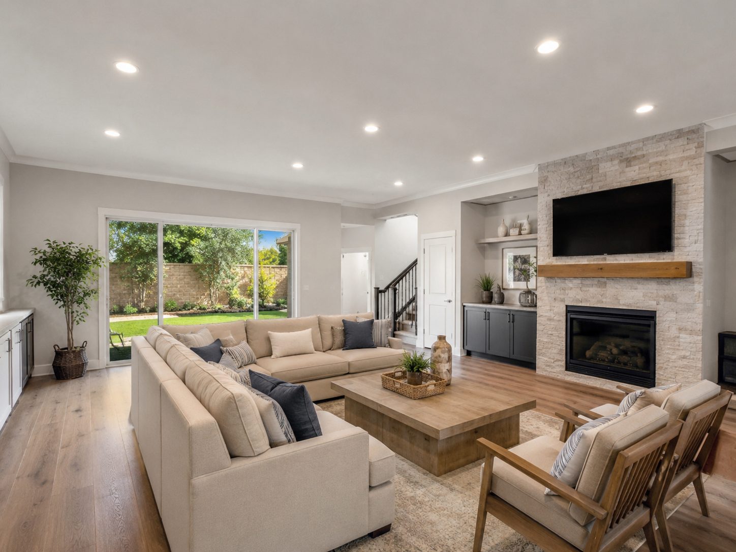 Living room with a stacked stone fireplace and wood mantel, wall mounted television, sectional sofa, and a sliding glass door onto the back lawn