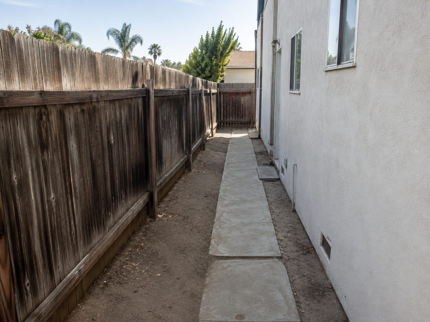 Narrow side yard of bare dirt beside a single concrete path, with a grey weathered wood fence